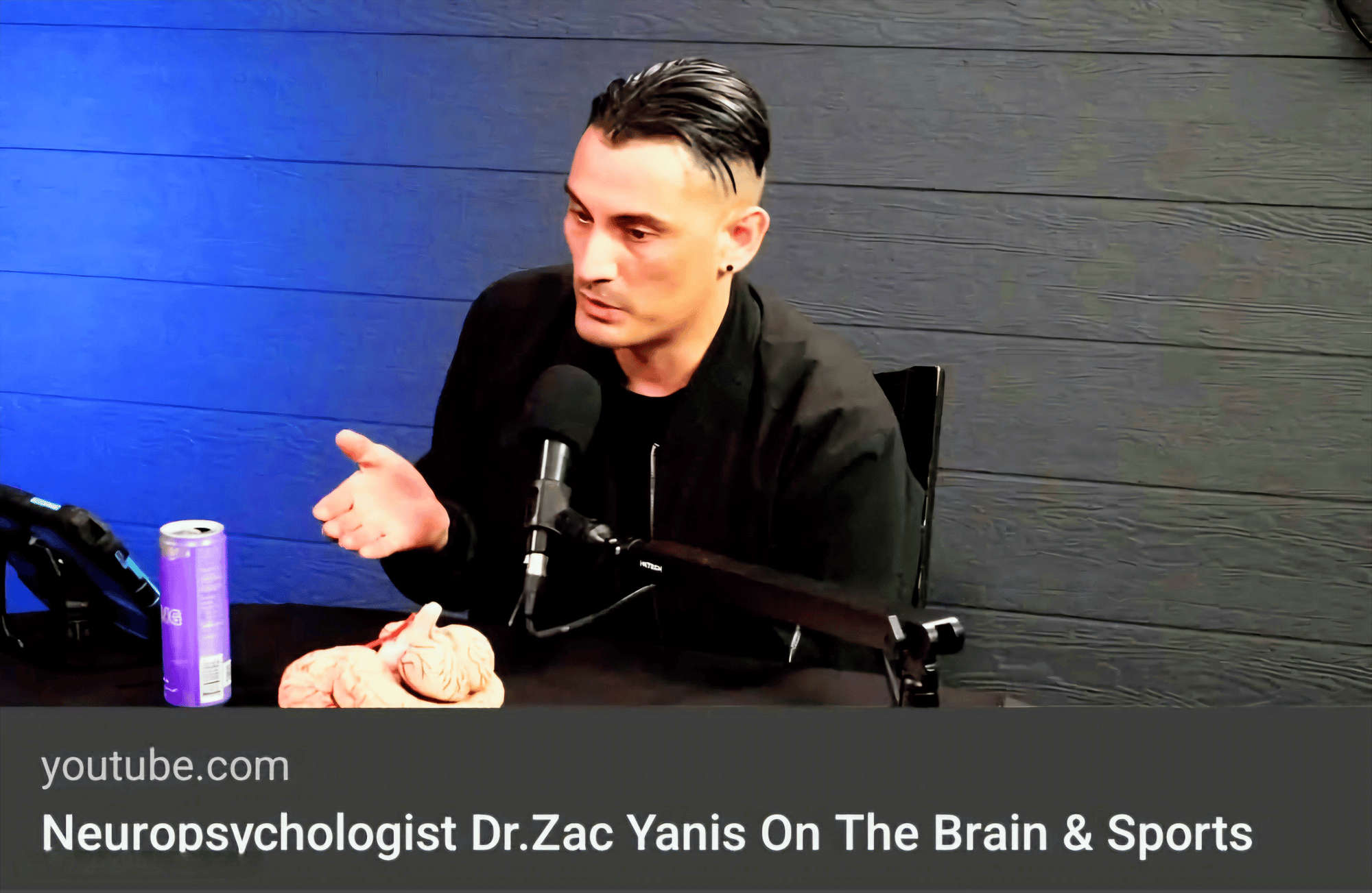 Dr. Zac speaking into a microphone during a video podcast in Atlanta, Georgia, discussing the brain and sports, with a brain model and energy drink on the table.