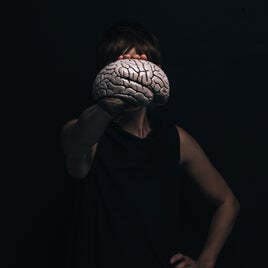 Person holding a realistic brain model in front of their face against a dark background, symbolizing neuroscience, psychology, and human performance.